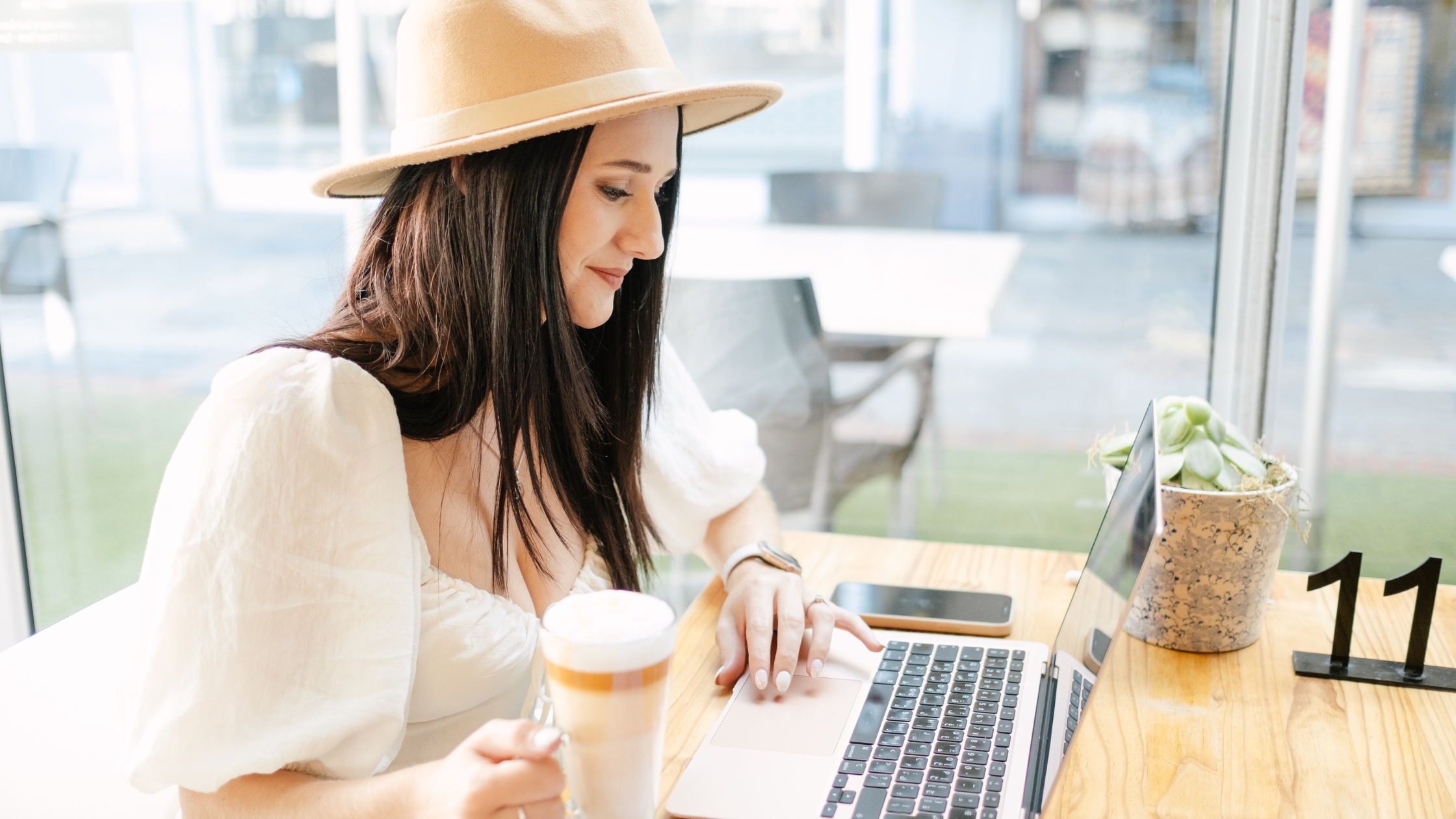 A woman wearing a stylish hat sits at a cafe table with a laptop, coffee, and plant nearby, busy creating automated email flows in Klaviyo following SheCommerce Simplified Tips