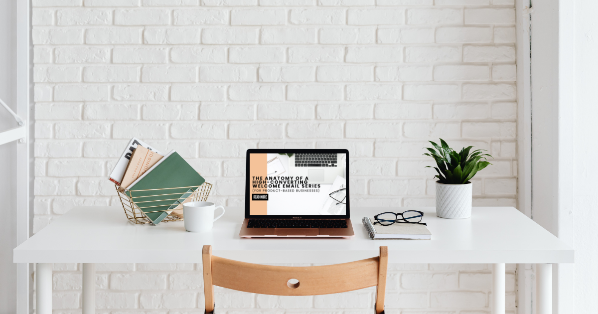 Minimalist workspace featuring a laptop displaying the title “The Anatomy of a High-Converting Welcome Email Series (For Product-Based Businesses),” surrounded by neatly arranged books, a plant, and glasses, on a white desk against a white brick wall.