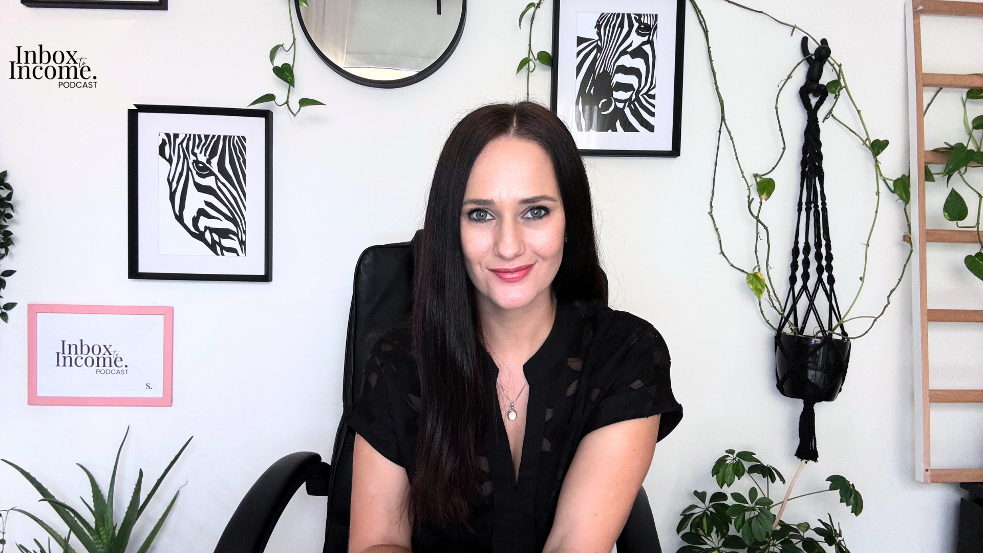 Liezle, host of the Inbox to Income Podcast, smiling while seated in a home office with framed artwork, plants, and a white wall background.